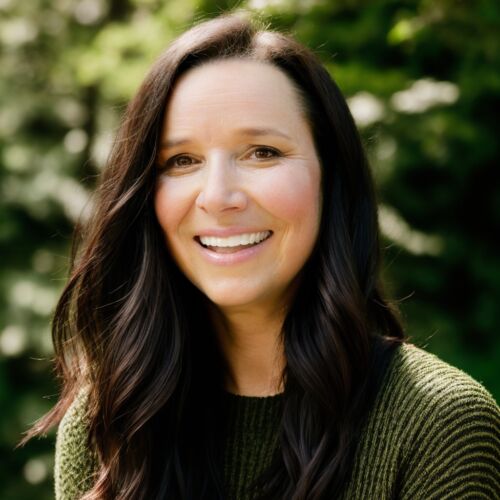 headshot of carrie hayes, she's wearing a green sweater with foliage in the background, looking at the camera and smiling