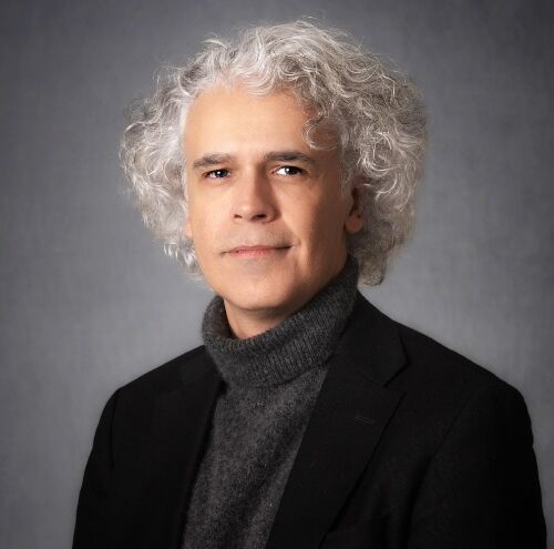 headshot of juan sanchez wearing a gray turtleneck and black blazer looking at the camera with a smirk