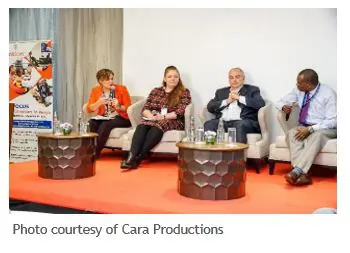 a conference panel with four individuals sitting in armchairs on a stage having a discussion